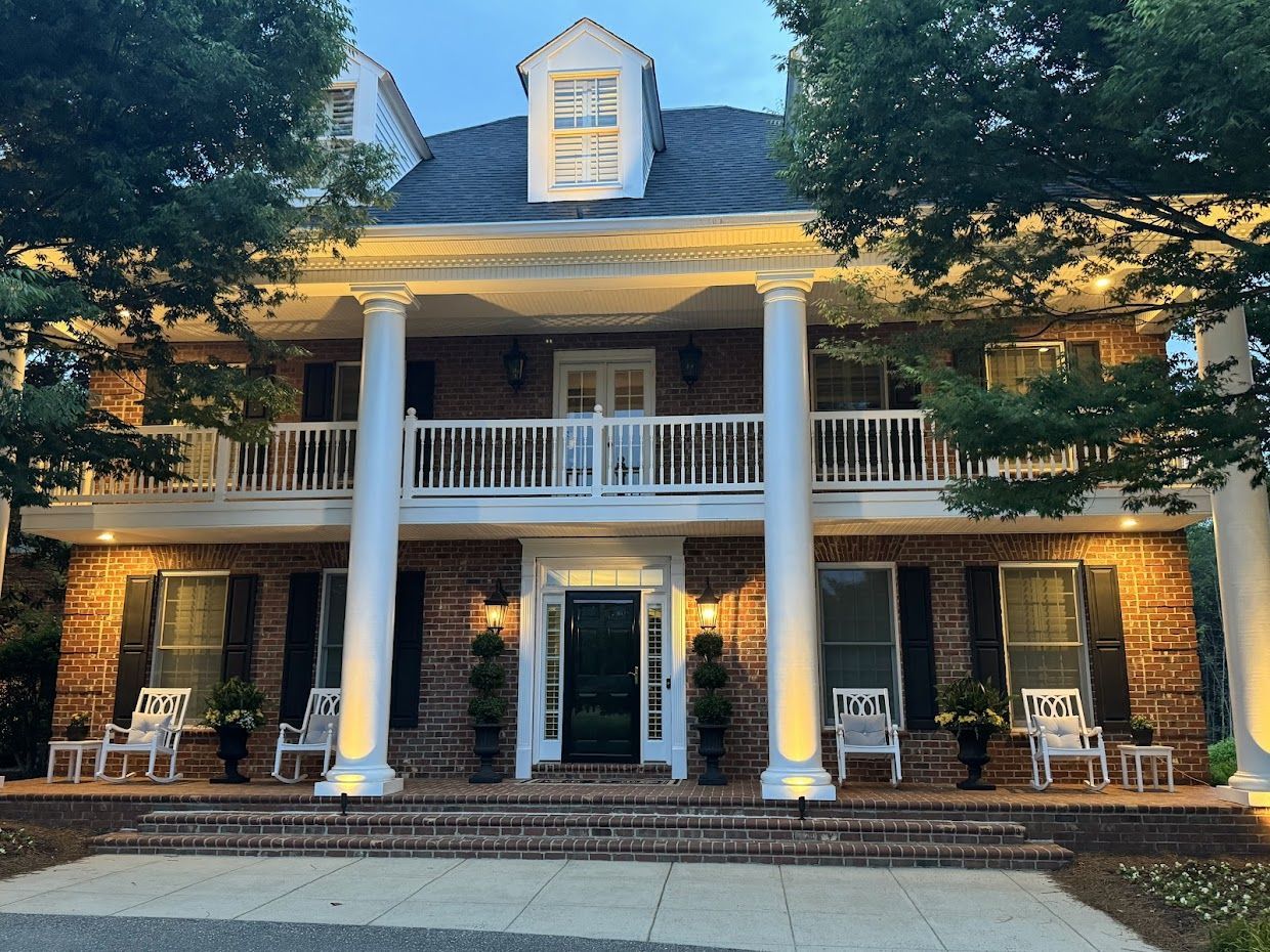 Two-story brick house with white columns and a balcony. Front porch with chairs and potted plants.