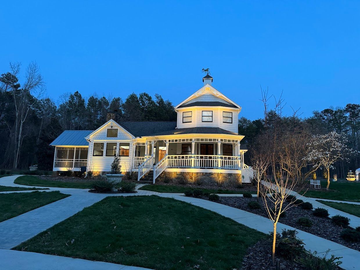 White house with tower, lit up at dusk, surrounded by landscaped grounds and trees.