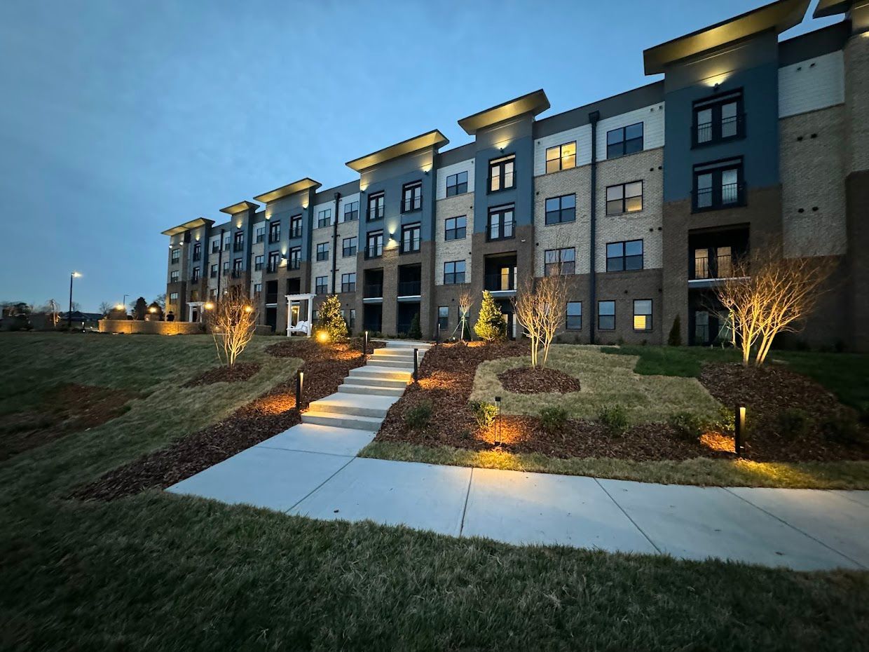 Apartment building exterior at dusk, with lit walkway and landscaping. Blue, gray, and tan facade.