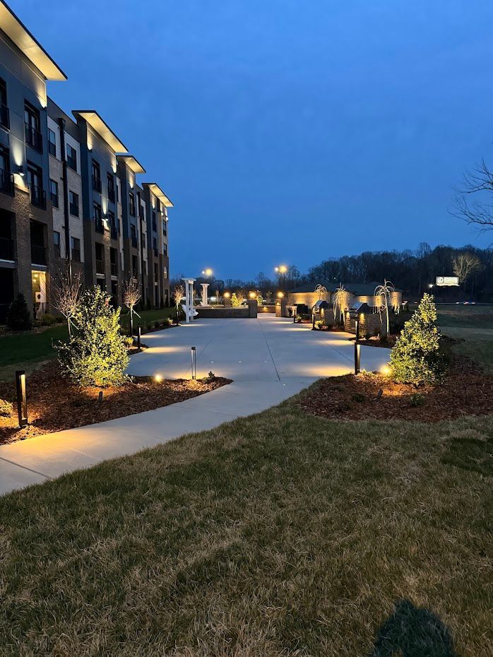 Exterior view of an apartment complex at dusk, illuminated by pathway lights, trees, and street lamps.