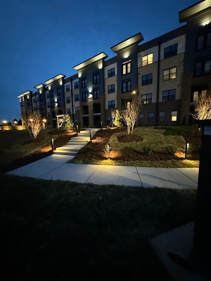 Apartment building exterior at night; illuminated path leading to entrance, landscaping with lights.