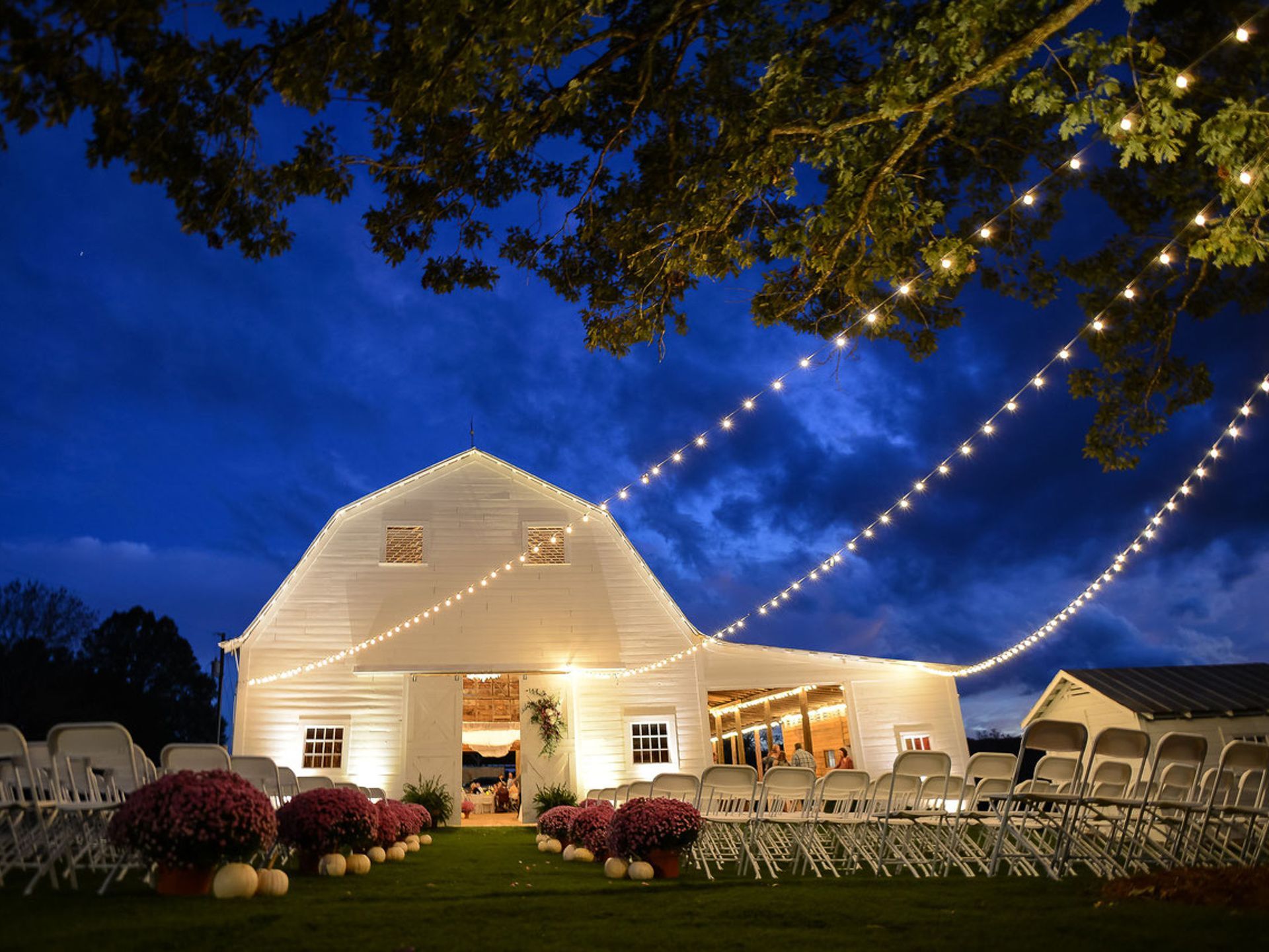 White barn illuminated with string lights, set up for an outdoor event with rows of chairs and potted plants.