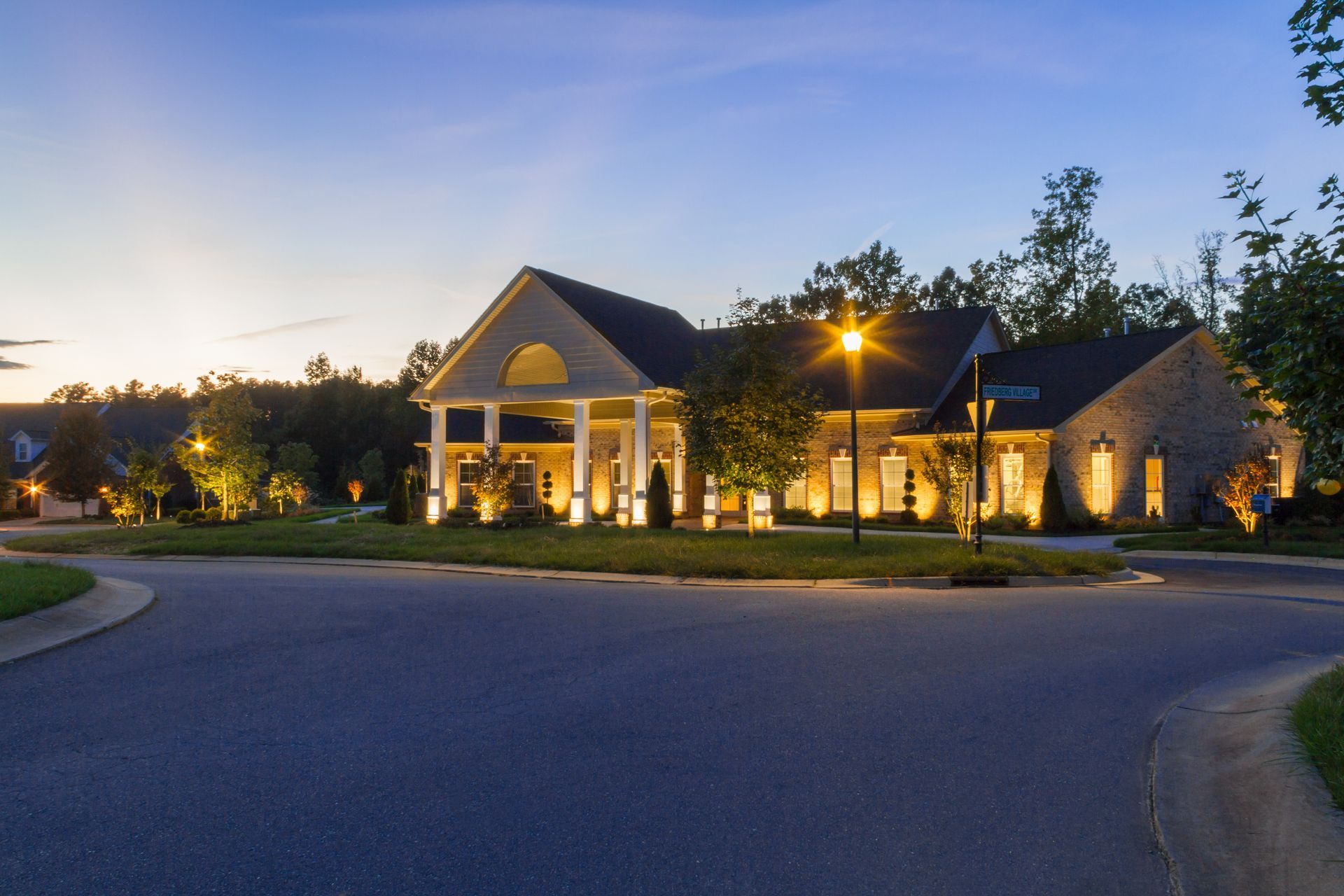 Suburban house at dusk, lit with warm exterior lights. A curved road leads to the property.
