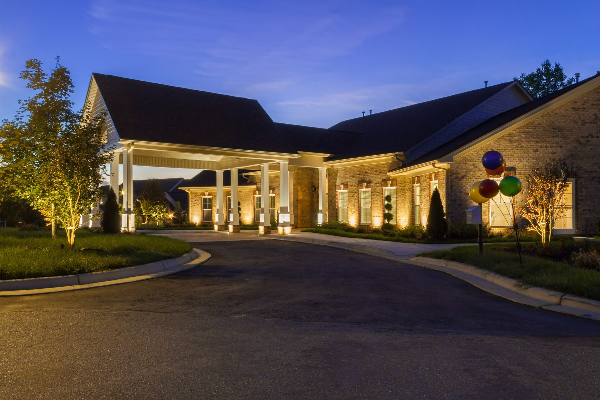 Exterior of a brick building with a covered entrance, lit up at dusk. Balloons on a post.