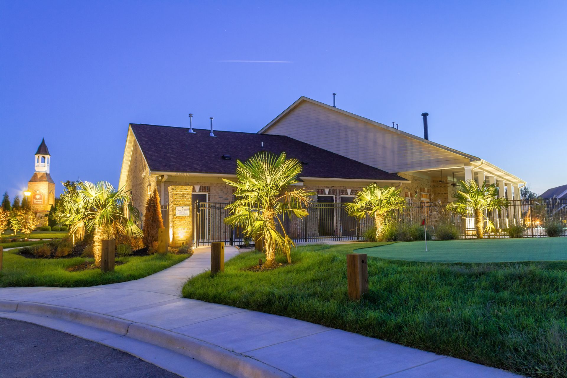 A modern building with a pathway and palm trees, lit at dusk. A tower is visible in the distance.
