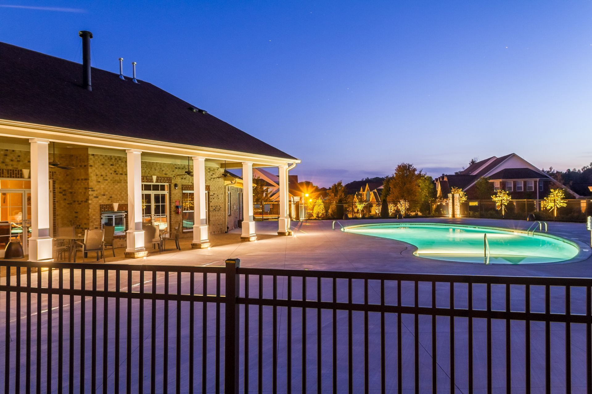 Pool and clubhouse at dusk; illuminated pool, brick building with white columns, black fence.