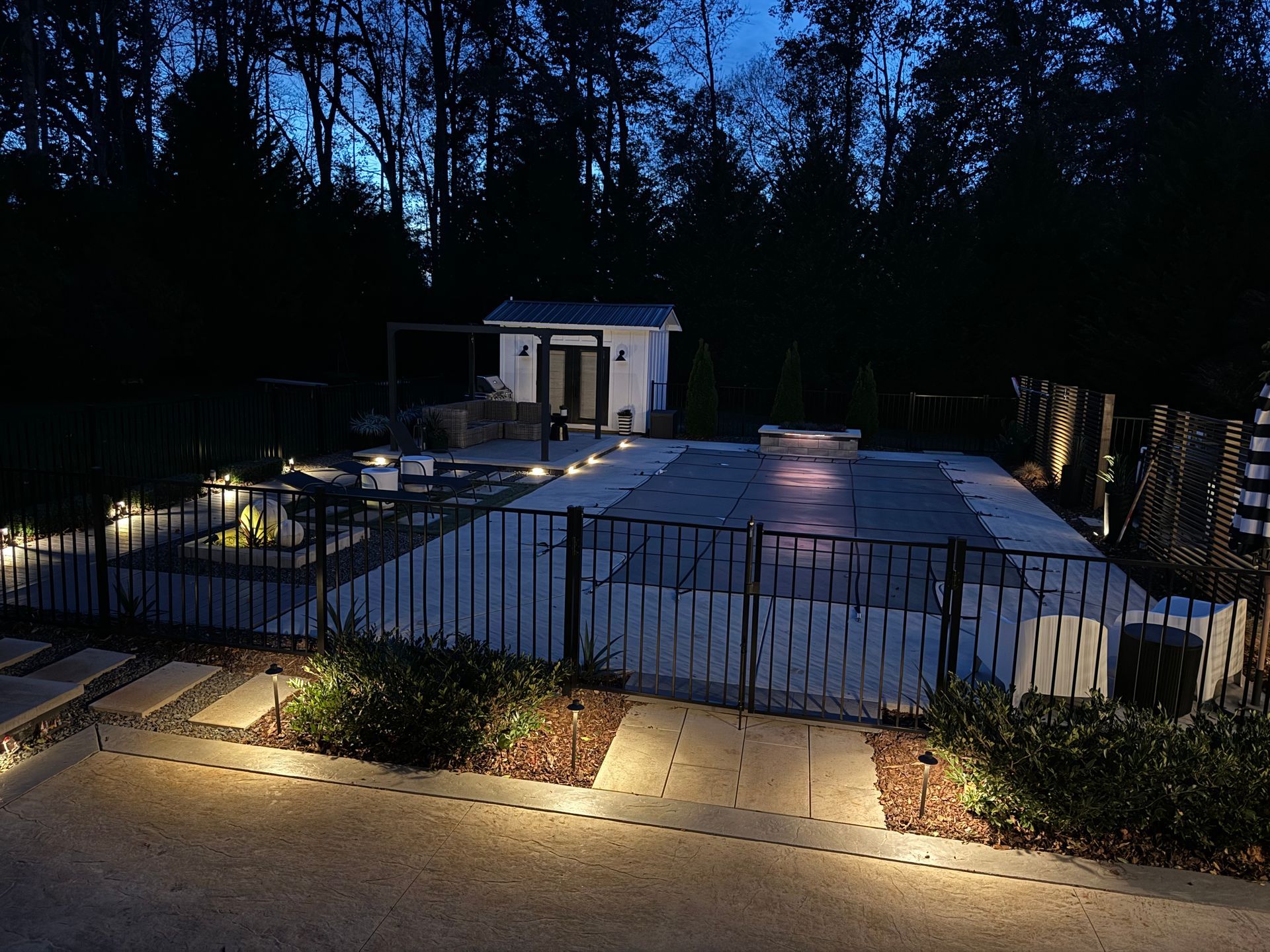 Nighttime view of a backyard with a pool, shed, and surrounding landscape illuminated by lights.