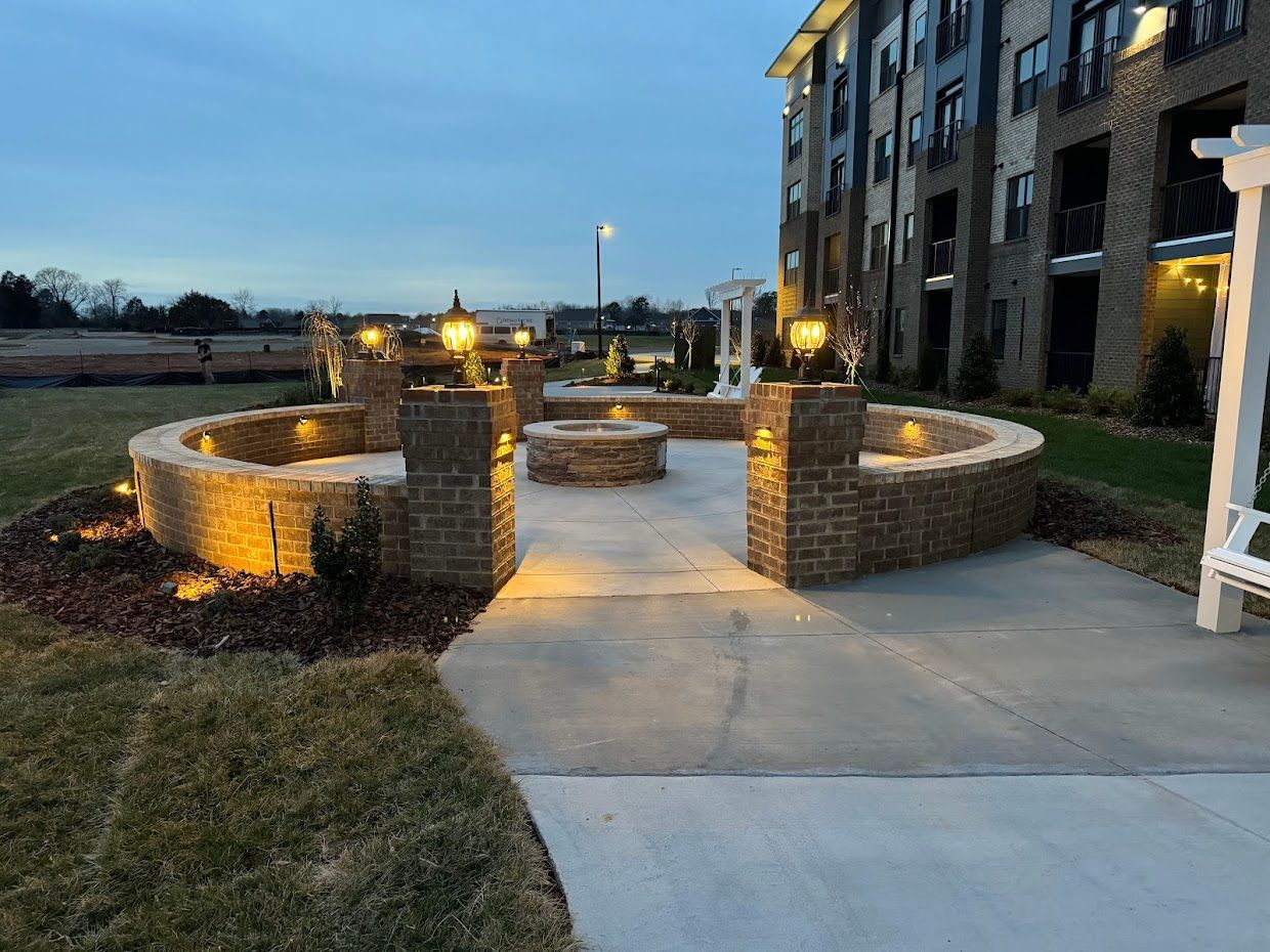 Outdoor seating area with brick walls, fire pit, and street lamps at dusk. Apartment building in background.
