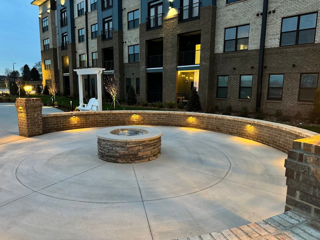 A brick fire pit in a circular concrete patio area, surrounded by a low brick wall and a multi-story building.