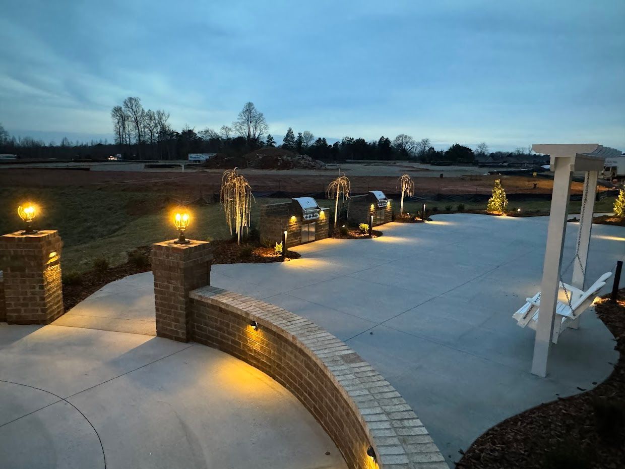 Concrete patio with brick pillars and built-in lighting, leading to a lawn under a twilight sky. A white swing is on the right.