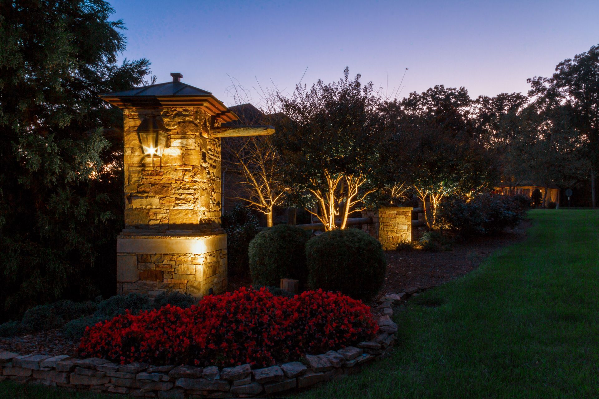 Stone entrance at dusk illuminated by warm lights, with red flowers and manicured bushes.