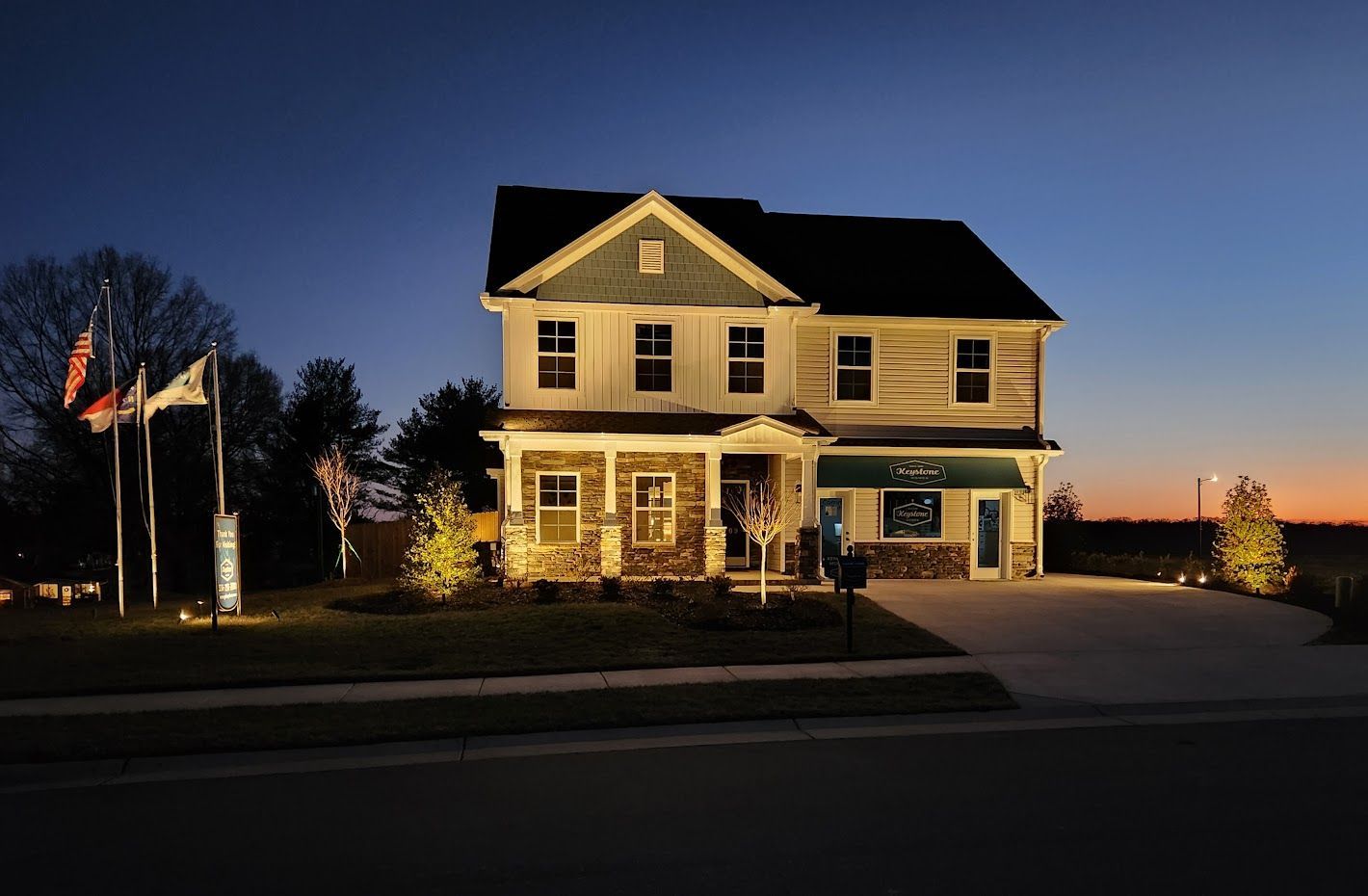 Two-story house lit up at dusk, with landscaping and flags in front. Blue and orange sky in the background.
