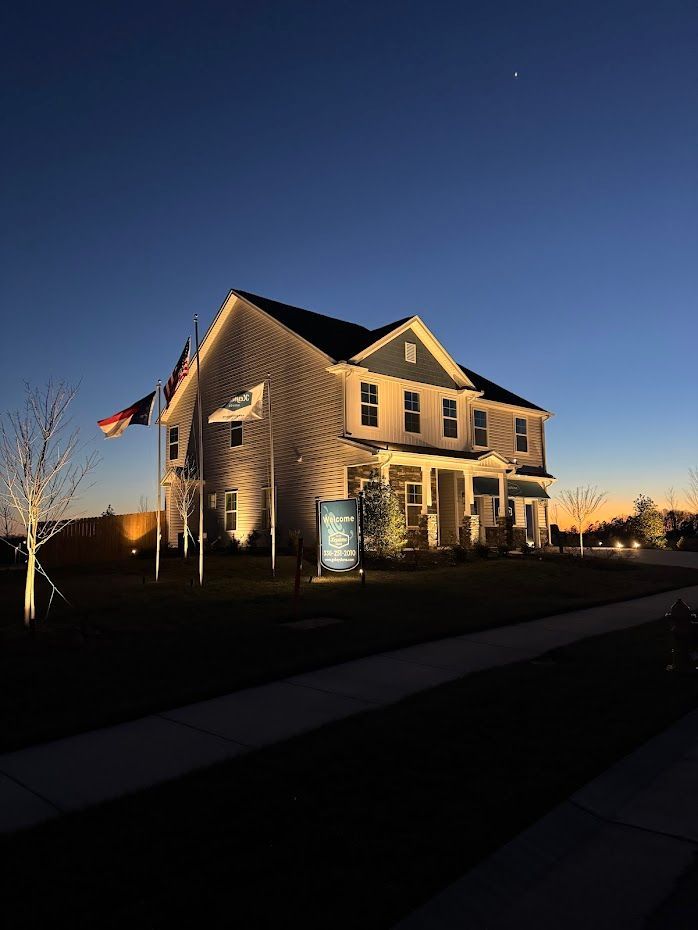 Two-story house at dusk, lit by spotlights. Flags and a sign in the front yard; blue and orange sky.