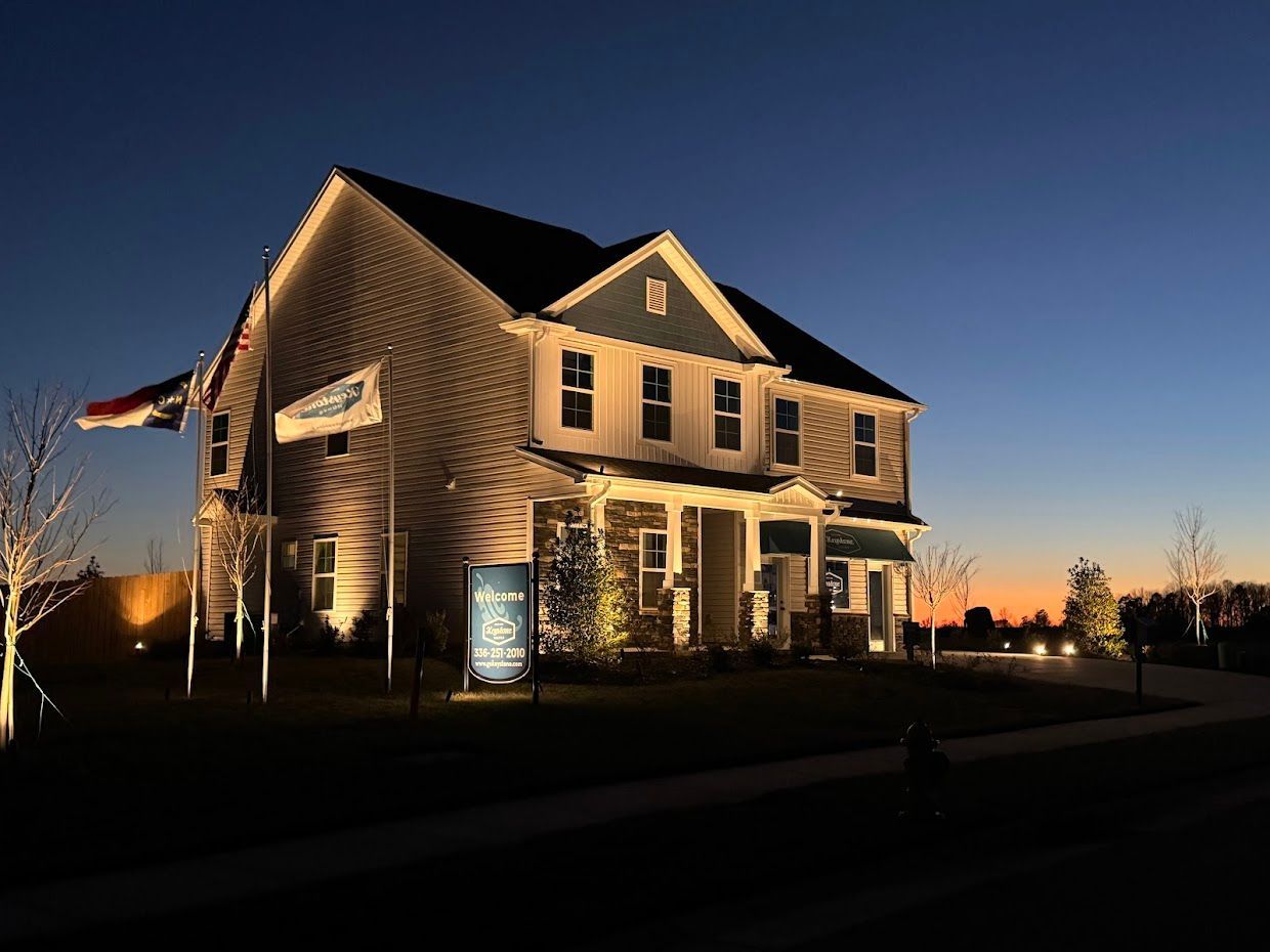 Two-story house lit at dusk with a blue and orange sky, a sign out front, and a flag.