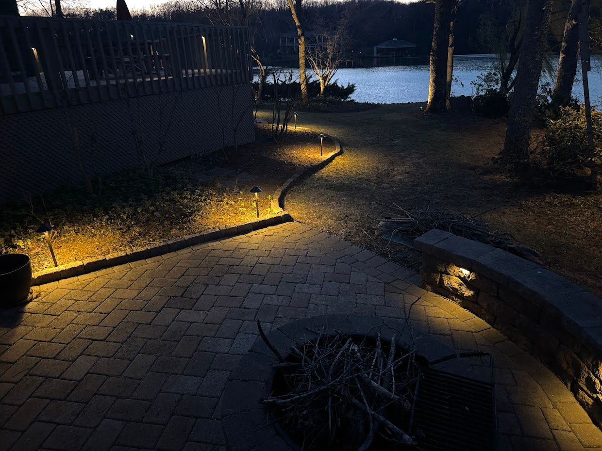 A brick pathway illuminated by landscape lights leads to a lake at dusk.