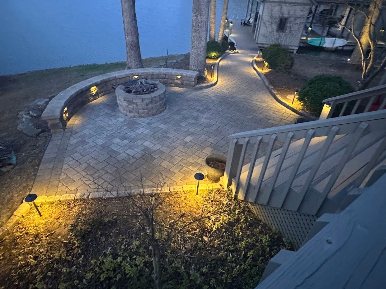 Stone patio with fire pit and pathway, lit with warm lights, by a lake.