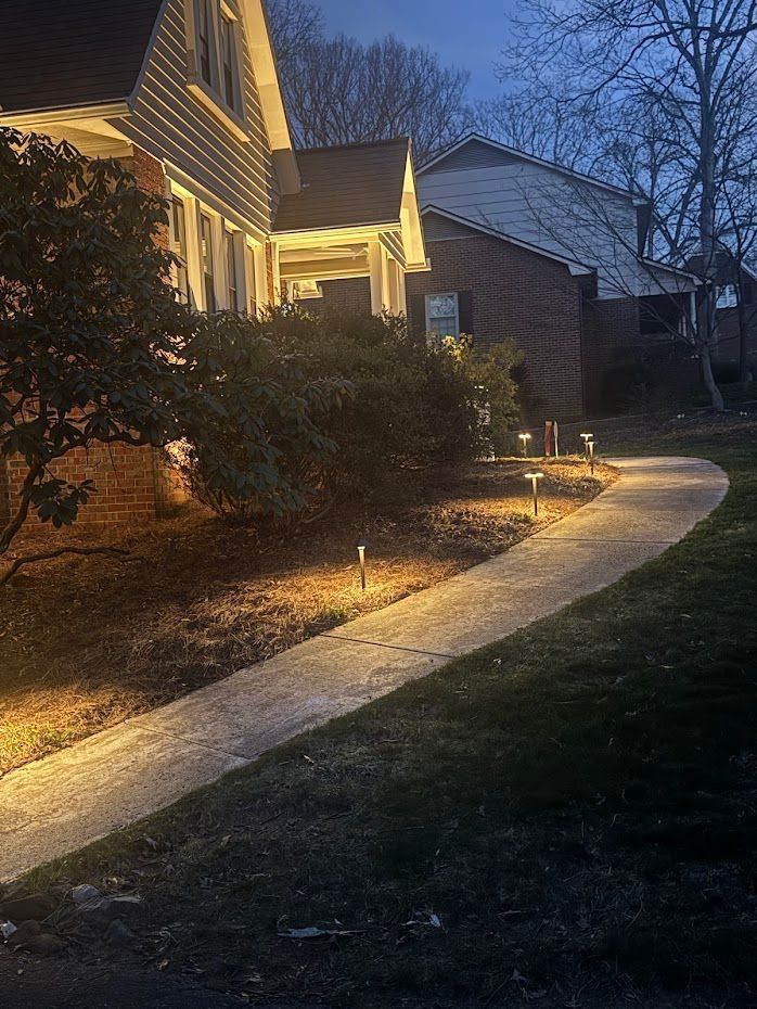 A paved walkway illuminated by outdoor lights leads to a house at dusk.