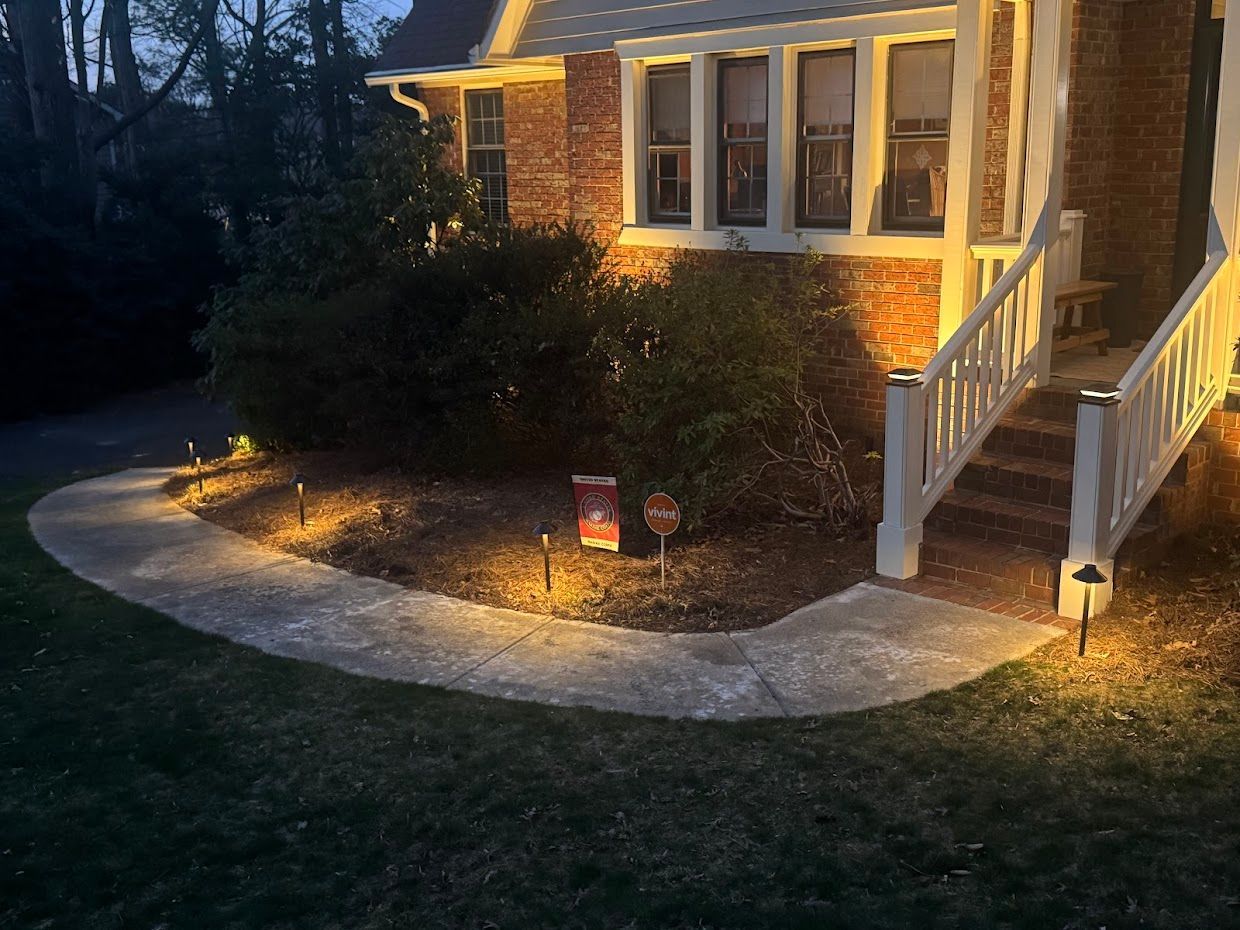 Pathway lit by spotlights leads to a house with a white railing and brick facade at dusk.