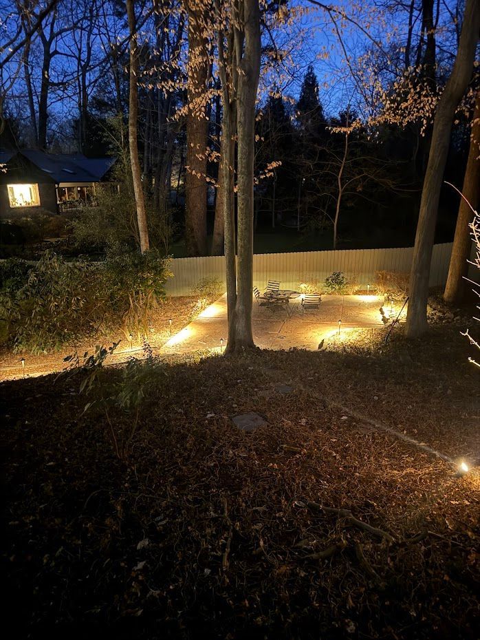 Nighttime view of a wooded area with trees and pathway illuminated by warm lights. House in background.