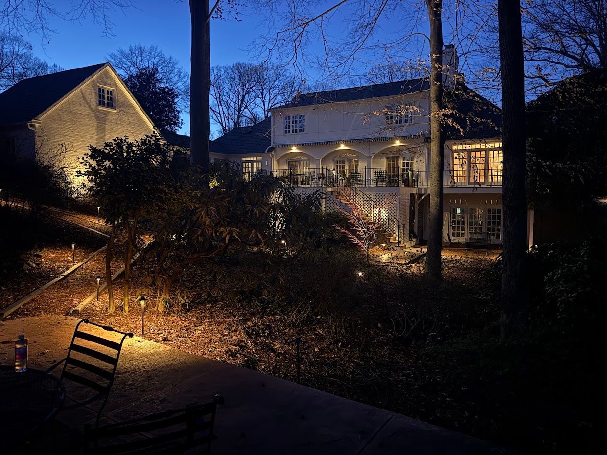 Exterior view of a large house at dusk with outdoor lighting illuminating the facade and trees.