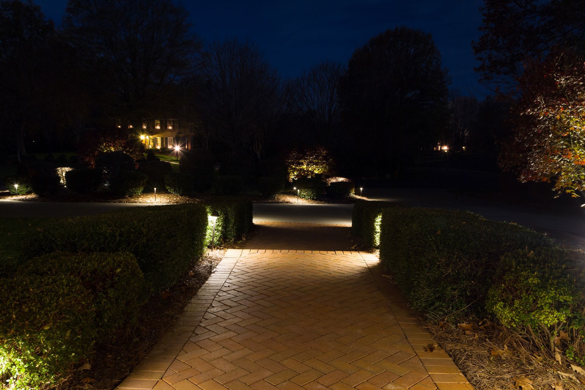 Brick path illuminated by warm lights, leading through hedges at night.