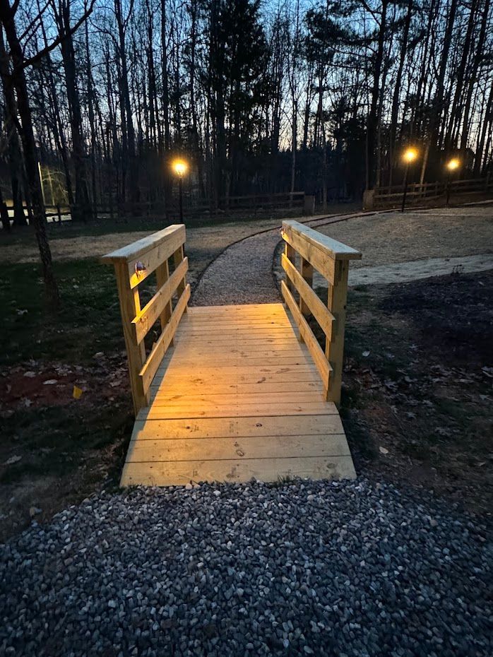 Wooden bridge over a gravel path in a wooded area, lit by streetlights.