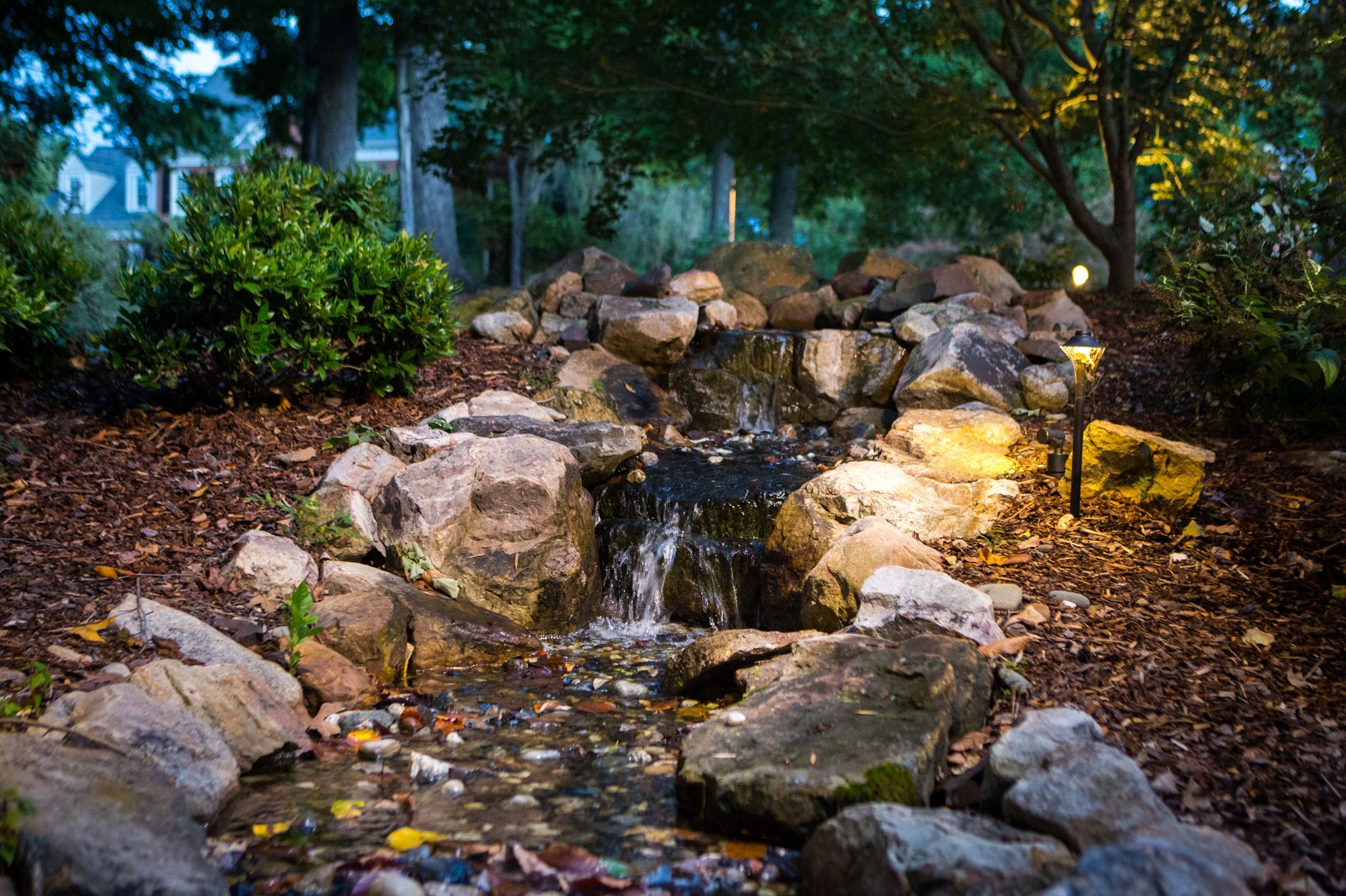 Small backyard waterfall cascading over rocks at dusk, lit by landscape lights.
