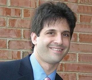 Man with dark hair smiles in front of a brick wall, wearing a suit and tie.