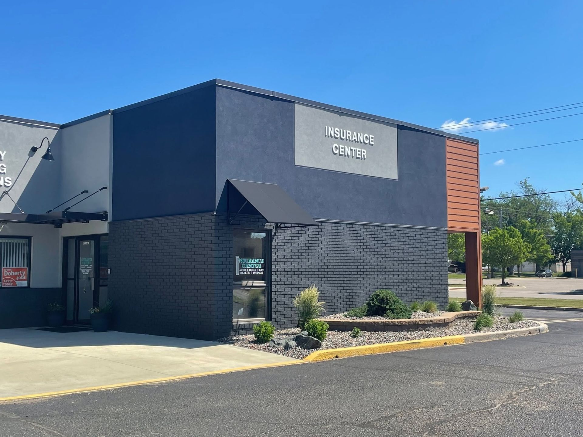 Modern commercial building exterior with dark gray facade, brick accent, and parking lot under clear blue sky