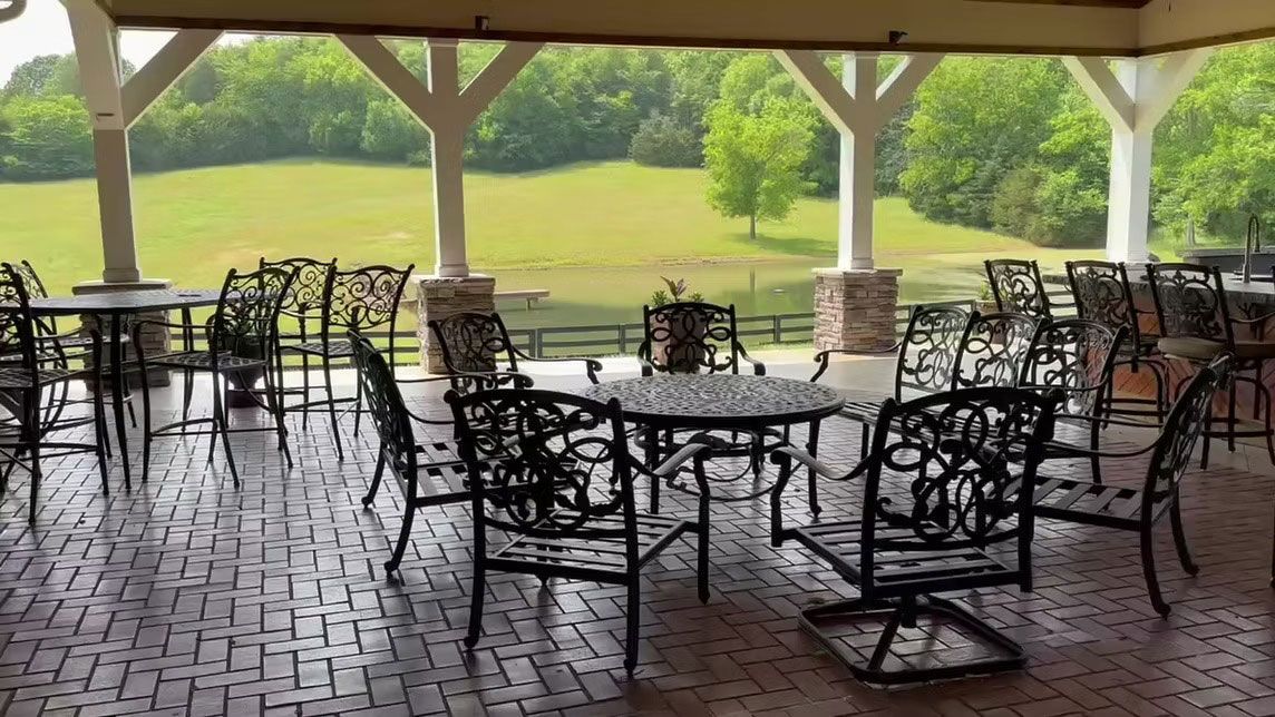 A covered brick patio with black metal tables and chairs overlooking a green field and pond.