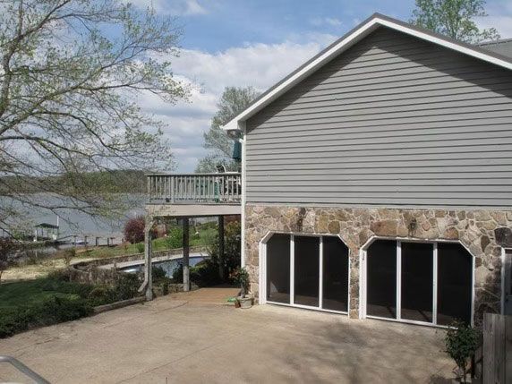 A gray-sided house with a stone foundation and two glass-paned garage doors, featuring a deck overlooking a lake.