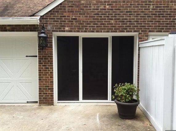 A brick home exterior features a white-framed triple screen door next to a white garage door and a potted plant.