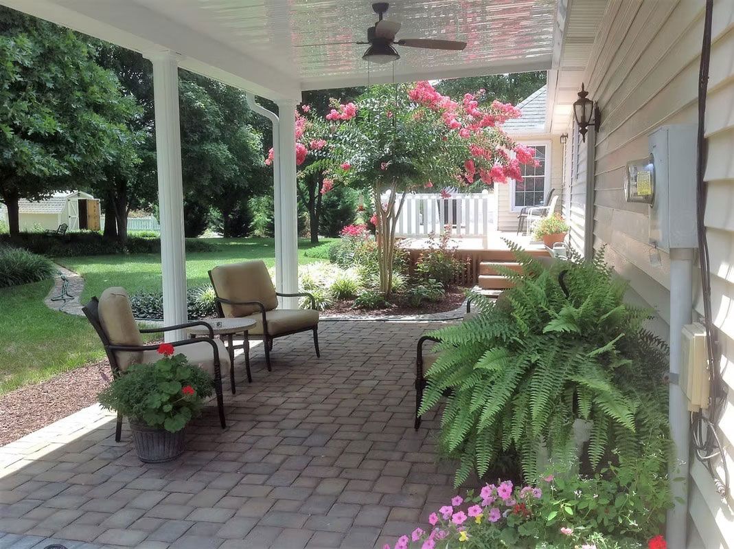 A covered brick patio with patio chairs, a ceiling fan, a large fern, and a flowering tree in a garden setting.