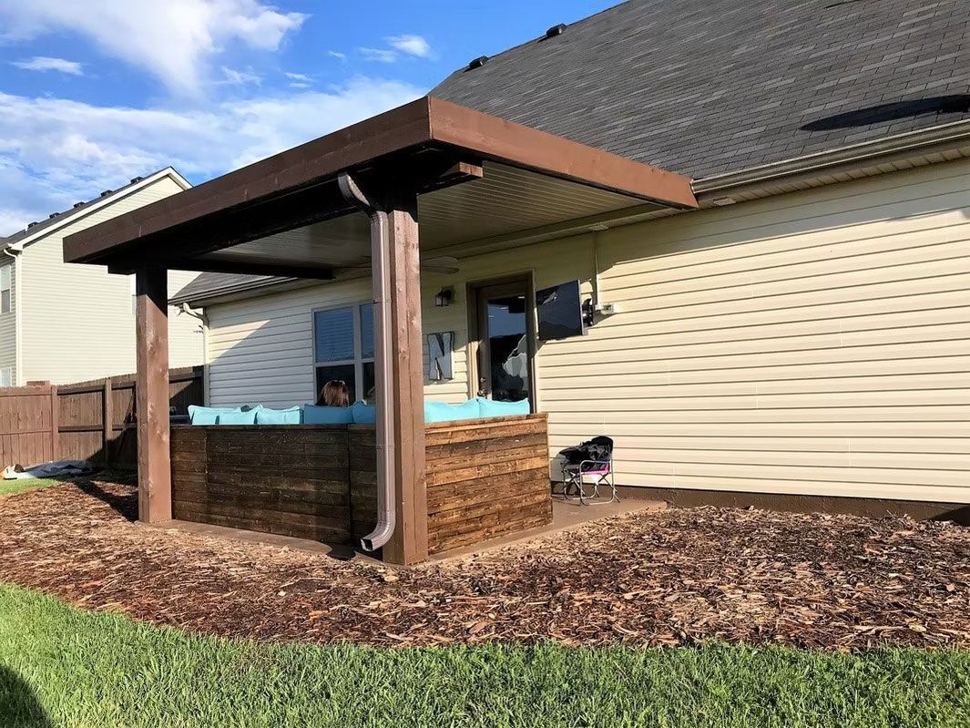 A wooden hot tub with light blue covers sits under a patio roof next to a house with beige vinyl siding and brown mulch.