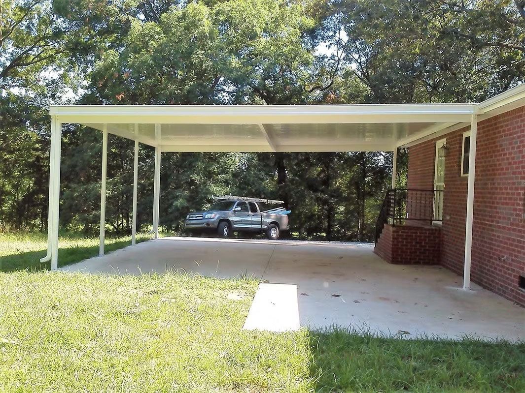 A white metal carport attached to a red brick house next to a concrete driveway, with a silver car parked in the shade.