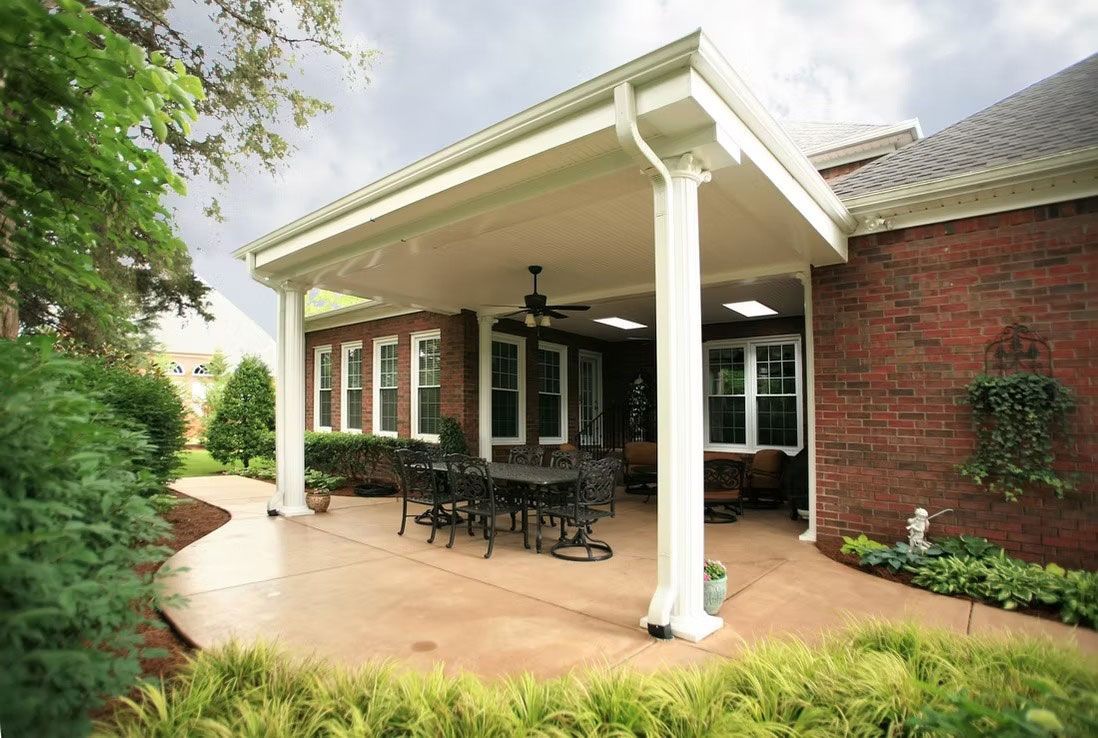 A brick house with a covered concrete patio featuring a dining set, ceiling fan, and white support columns.