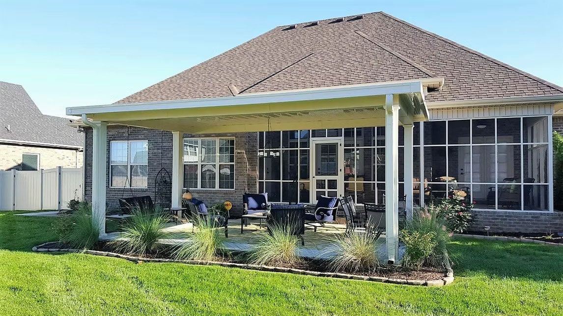 A backyard patio featuring a white pergola over a concrete floor, set against a brick house with an enclosed porch.