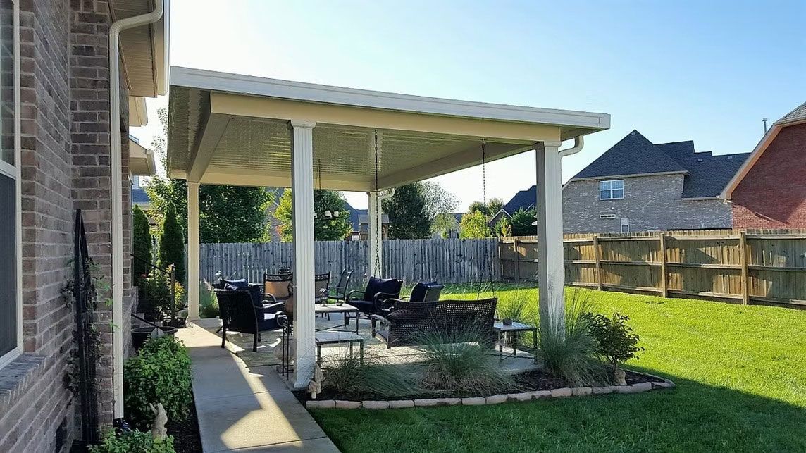 A patio with a light-colored roof cover, patio furniture, and plants set in a backyard with a wooden fence.