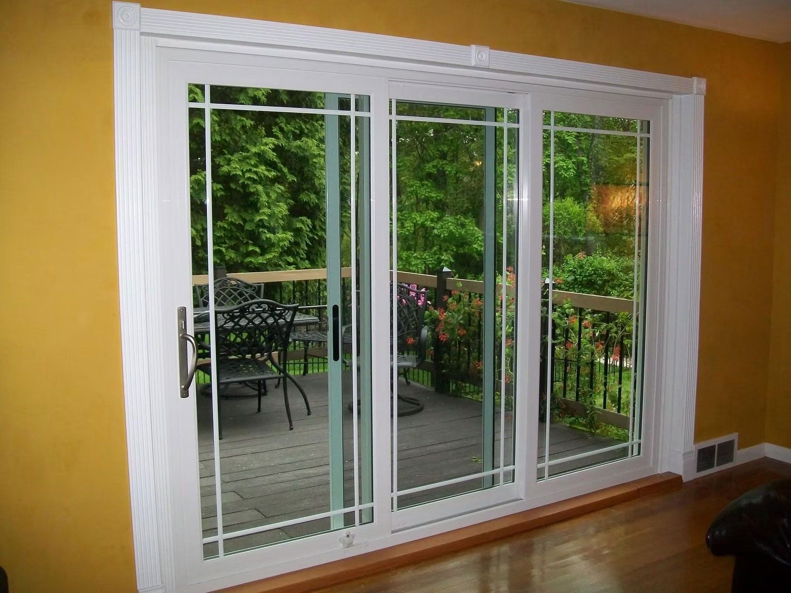White sliding glass patio doors with grid designs against a mustard yellow wall, opening to a wooden deck with furniture.