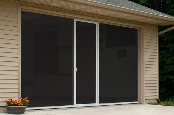 A white-framed sliding screen door installed on a garage opening, set against beige siding with a potted plant.