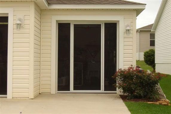 A white-trimmed screen door with two sidelights on a beige-sided house with a concrete porch and a small green bush.