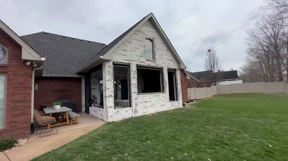 A residential brick house features a white stone-faced addition with empty window frames and a rear patio.