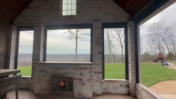 A screened-in porch with a brick fireplace, white washed walls, and views of a green lawn and trees in the distance.