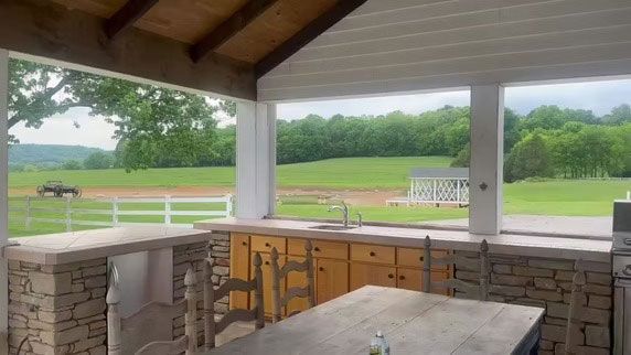 A stone-base outdoor kitchen with a sink under a covered patio, overlooking a grassy field and a distant white fence.