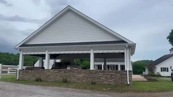 A white pavilion with a stone base and open sides sits on a lawn under a cloudy sky next to a matching white building.