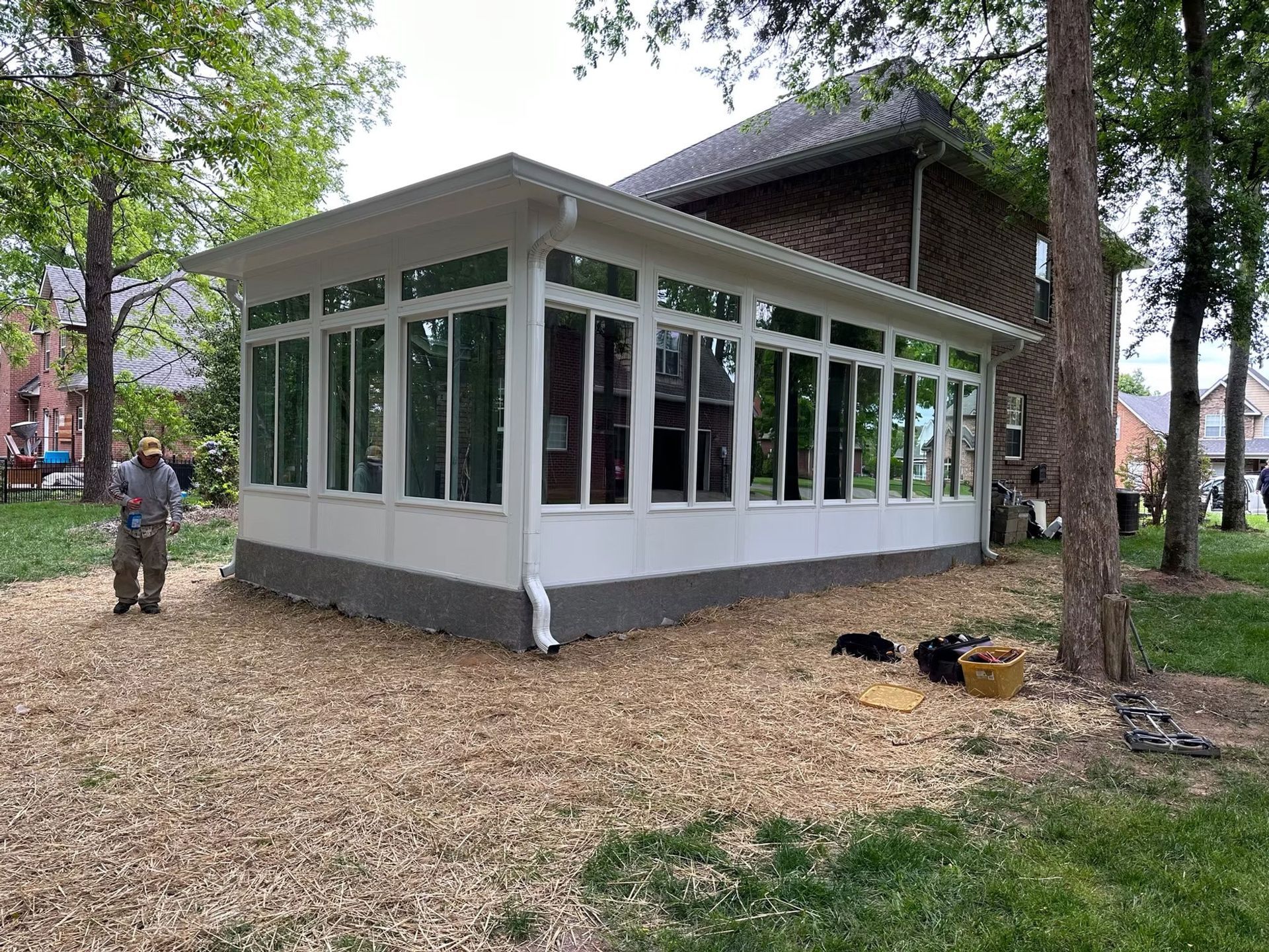 A white sunroom addition attached to a brick house stands in a yard covered with wood chips, with a person standing nearby.