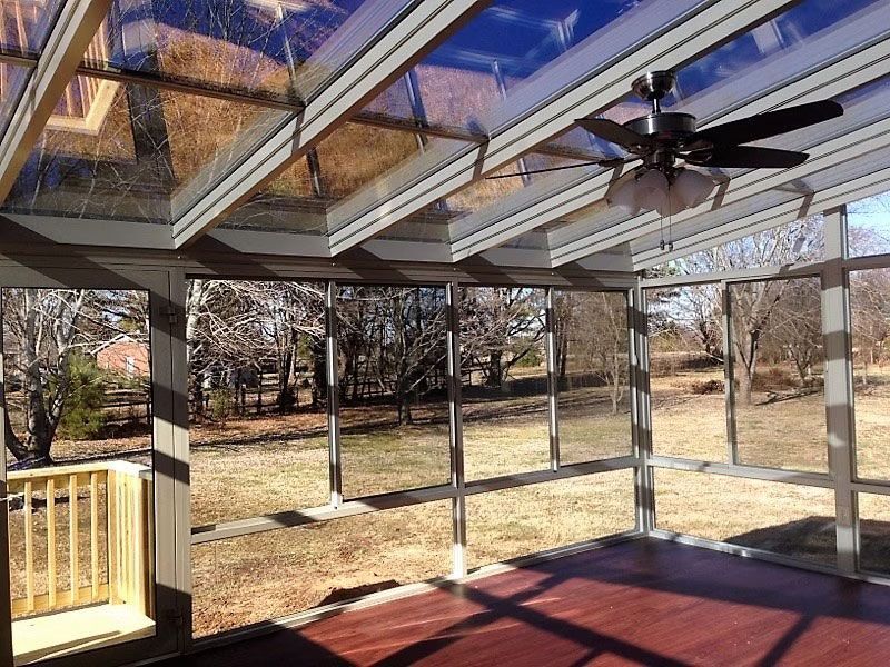 A view from inside a sunroom with a glass roof, a ceiling fan, and large windows overlooking a grassy yard with trees.
