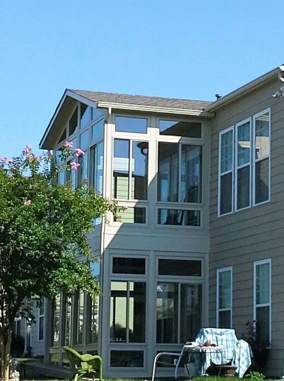 A two-story beige home with a sunroom extension, large windows, and a tree in the foreground.