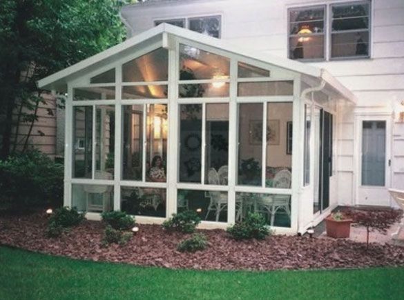 A white sunroom addition attached to the back of a house, featuring large glass windows and a mulch garden bed in front.