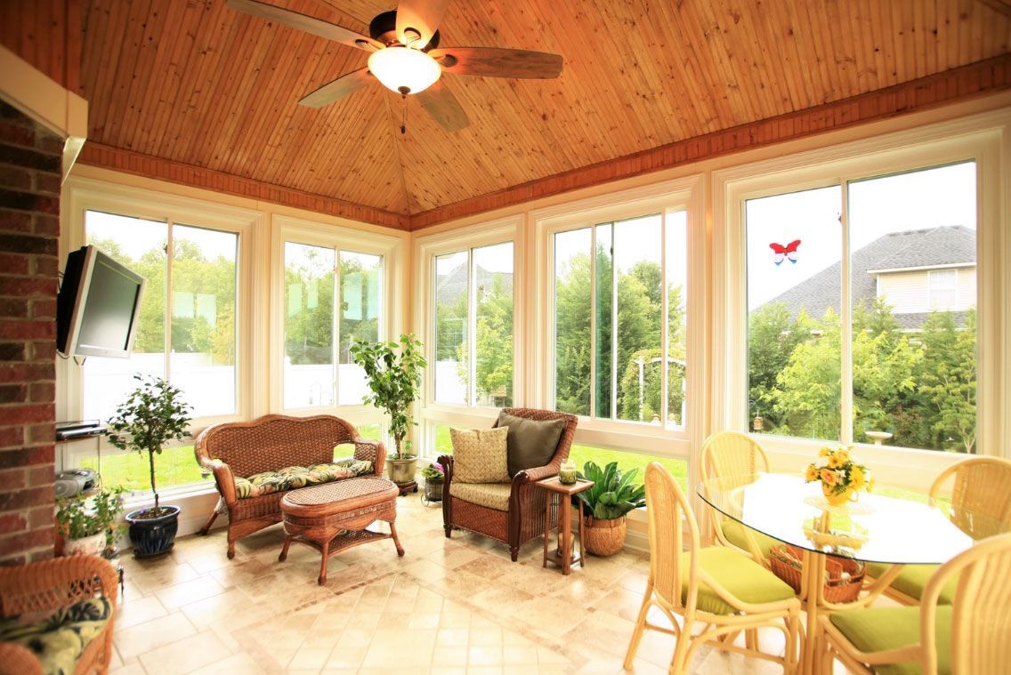 A sunroom with a wood-paneled ceiling, tiled floors, wicker furniture, and large windows overlooking a green landscape.